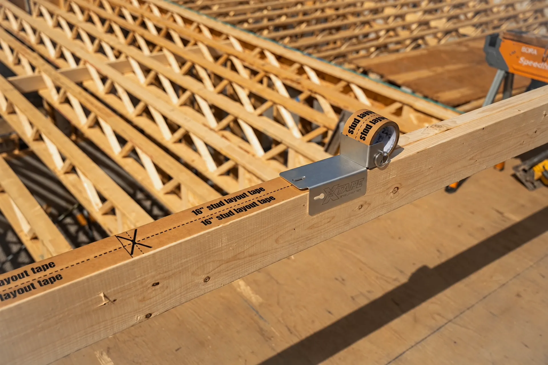 L’applicateur Stud Layout Tape sur un 2×4, son rouleau chargé, le ruban X-Tape déjà posé derrière lui, les poutrelles du plancher au fond